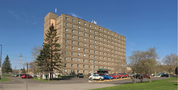 Ten-story apartment building with a parking lot out front and a blue sky behind.